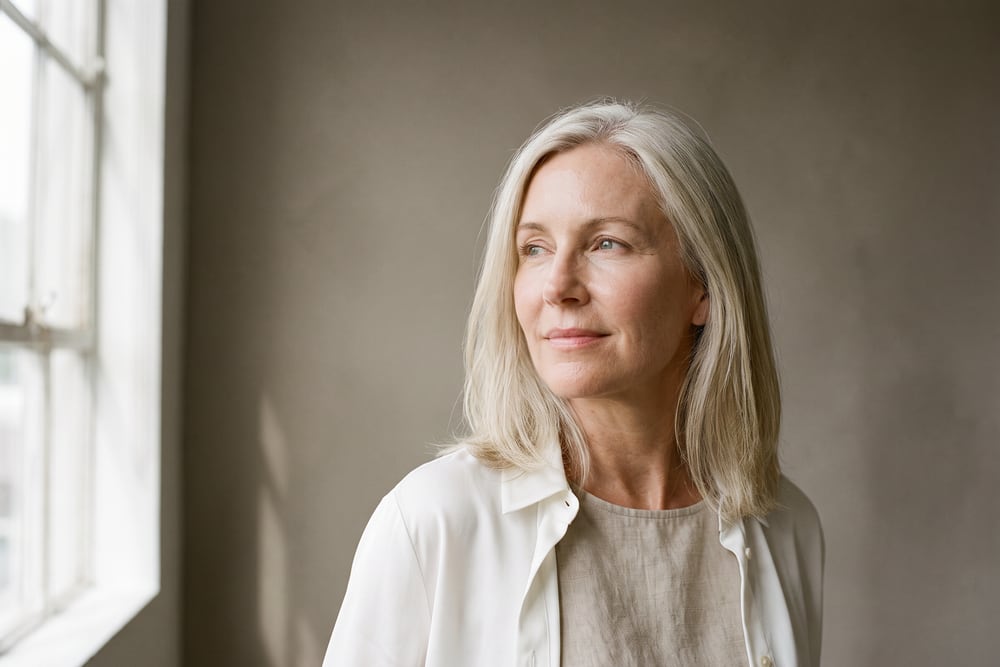 Woman in her fifties with silver-blonde hair looking toward a window, the kind of patient who asks about eyelid surgery in San Antonio